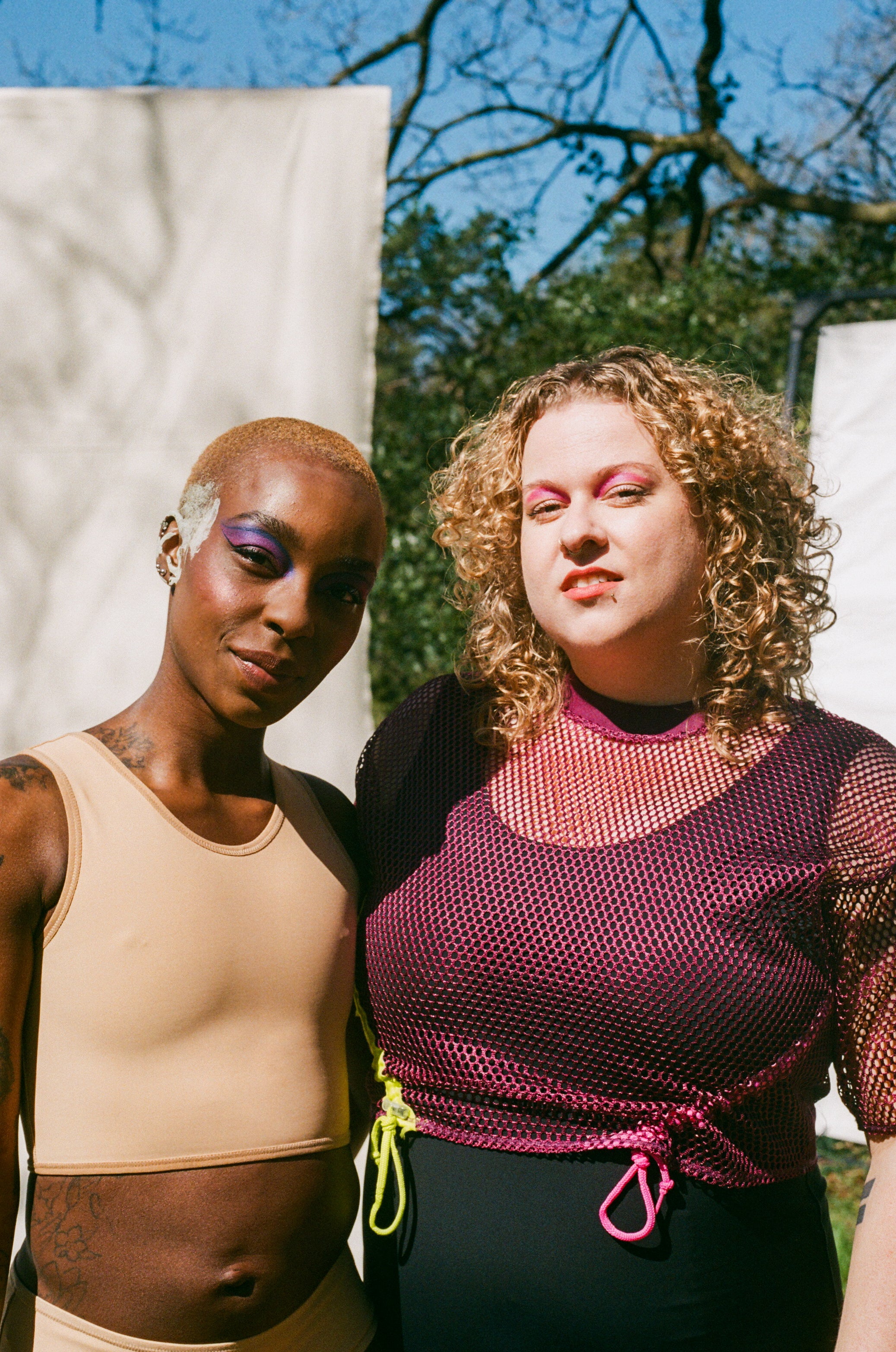 Our models Sylvia and Lavinia posing for a photo - lavinia wears a short binder and has purple eyeshadow. Sylvia wears pink eyeshadow and a trans bathing suit
