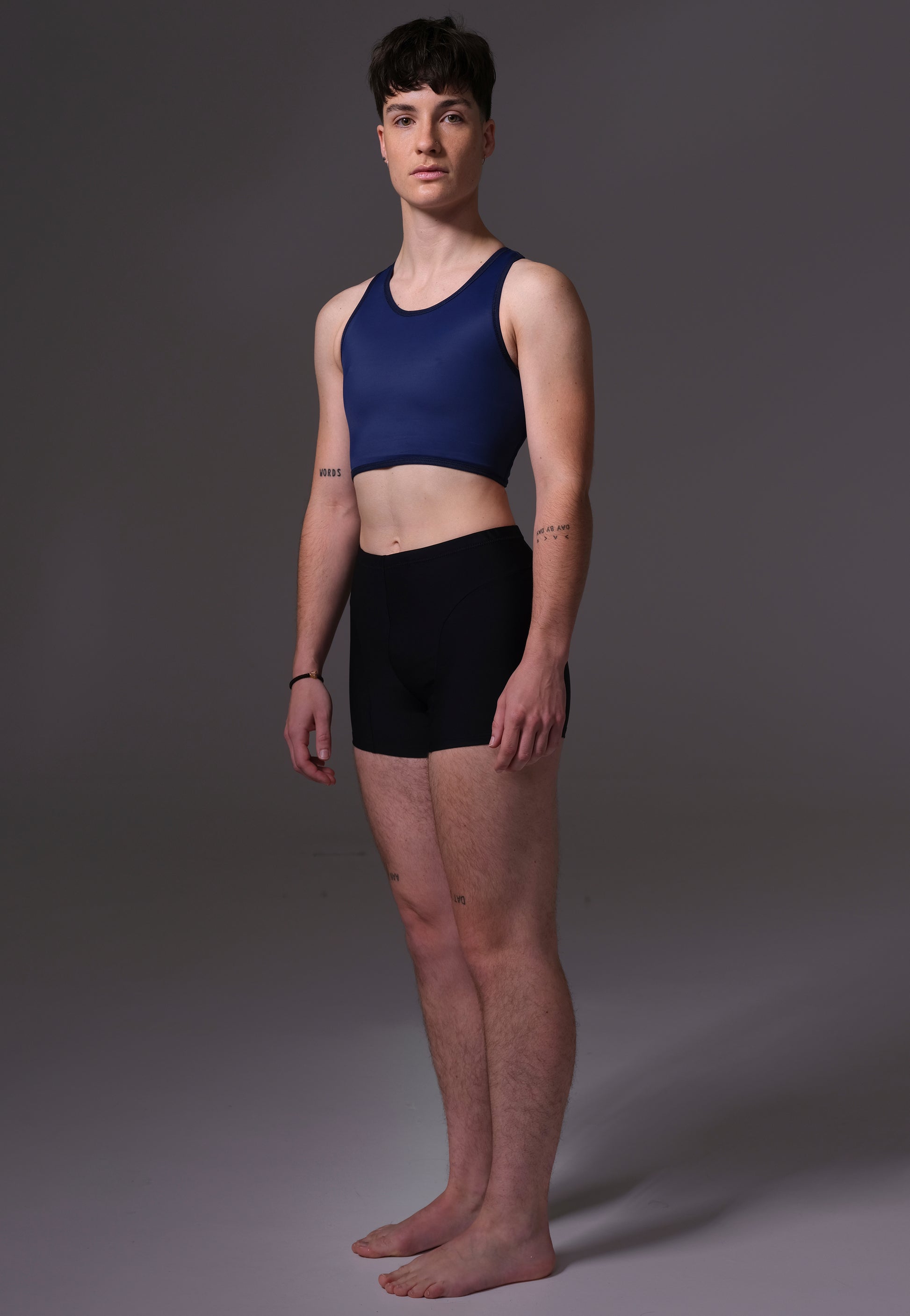 Non-Binary model Chris wearing a navy blue Tank Swim Binder and black shorts on a gray background