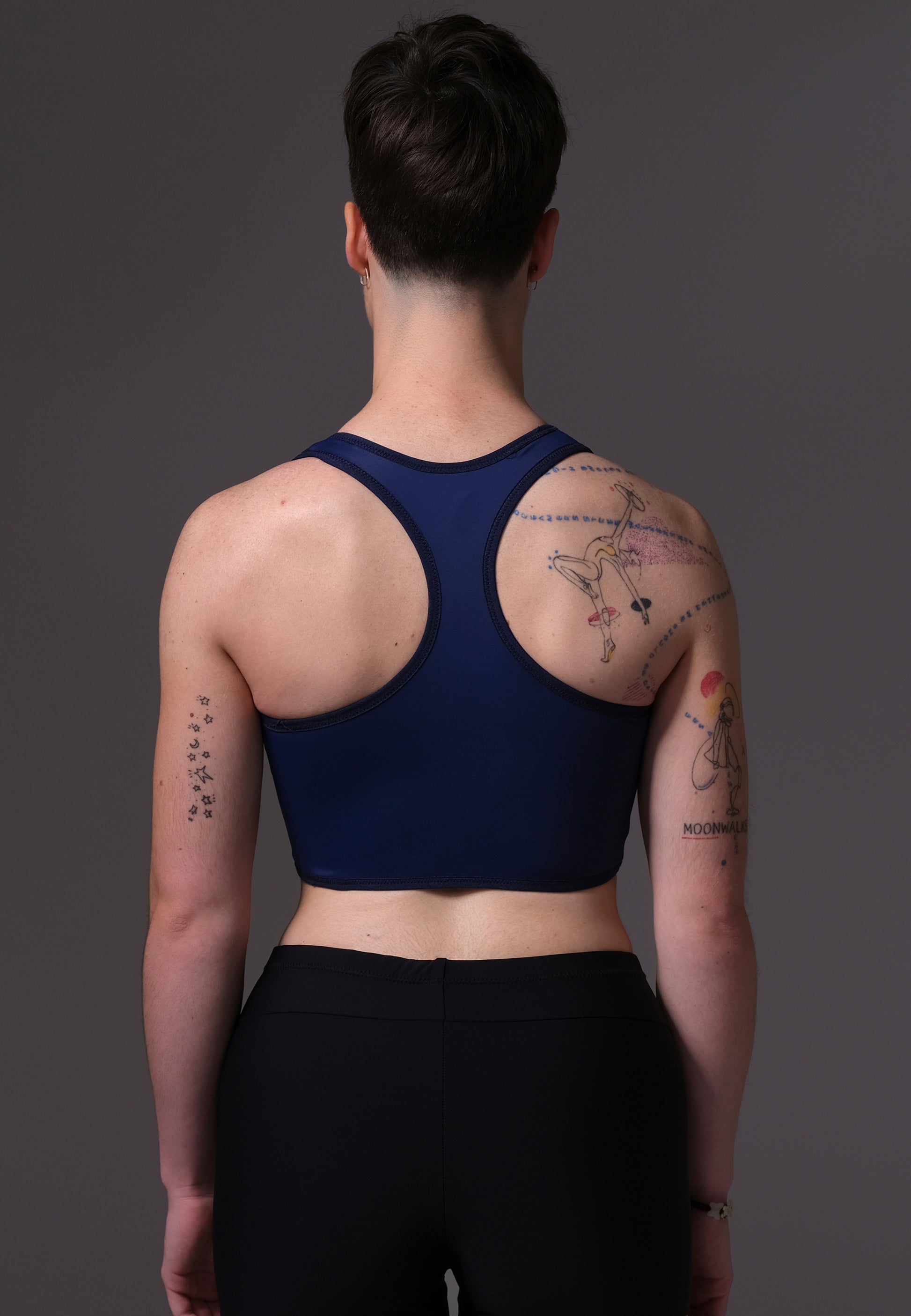 Non-Binary model Chris wearing a navy blue Tank Swim Binder and black shorts with tattoos on a dark background