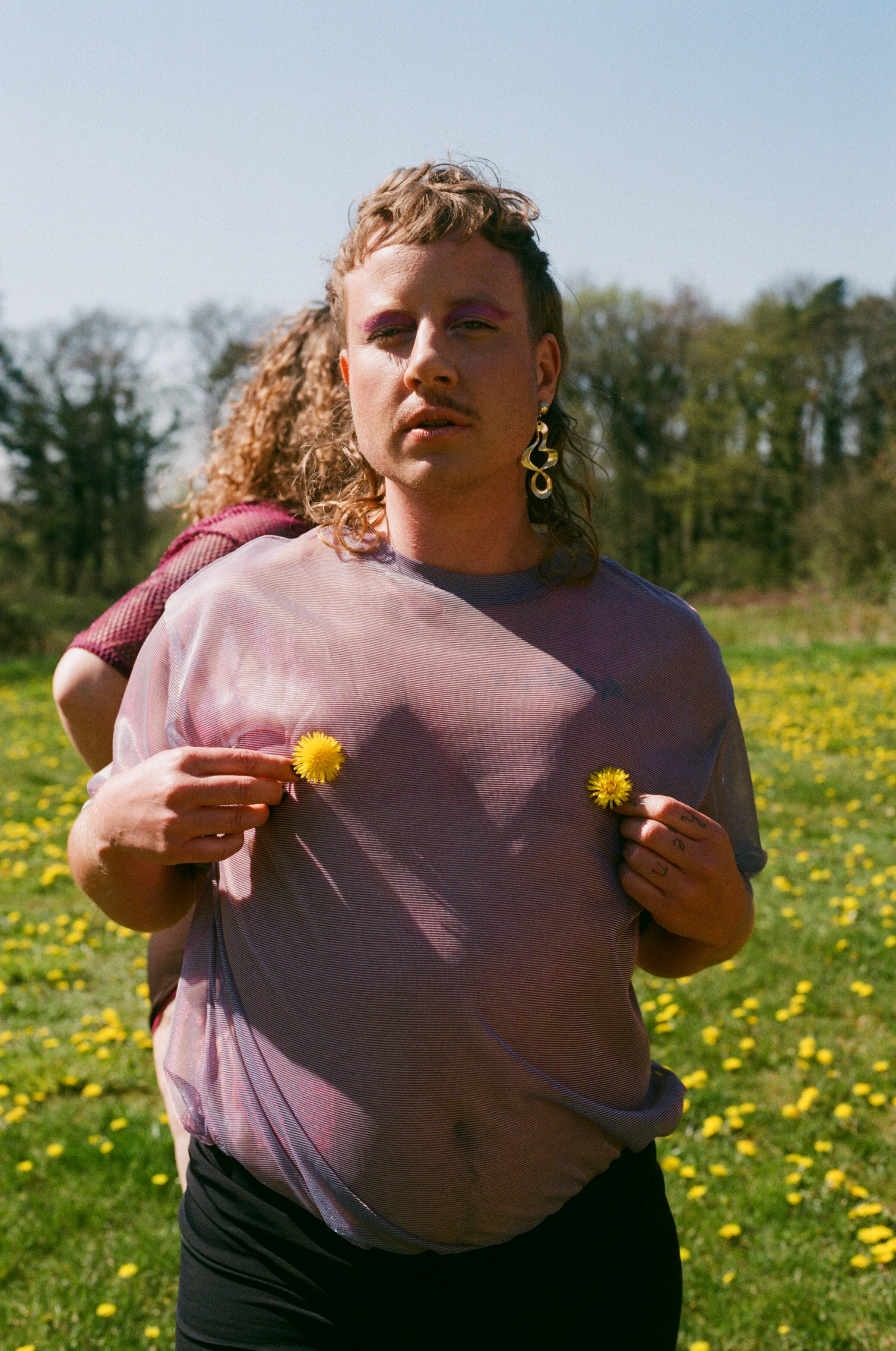 Our non-binary model Storm holds two yellow flowers and looks into the camera. You can faintly see his mastectomy scars
