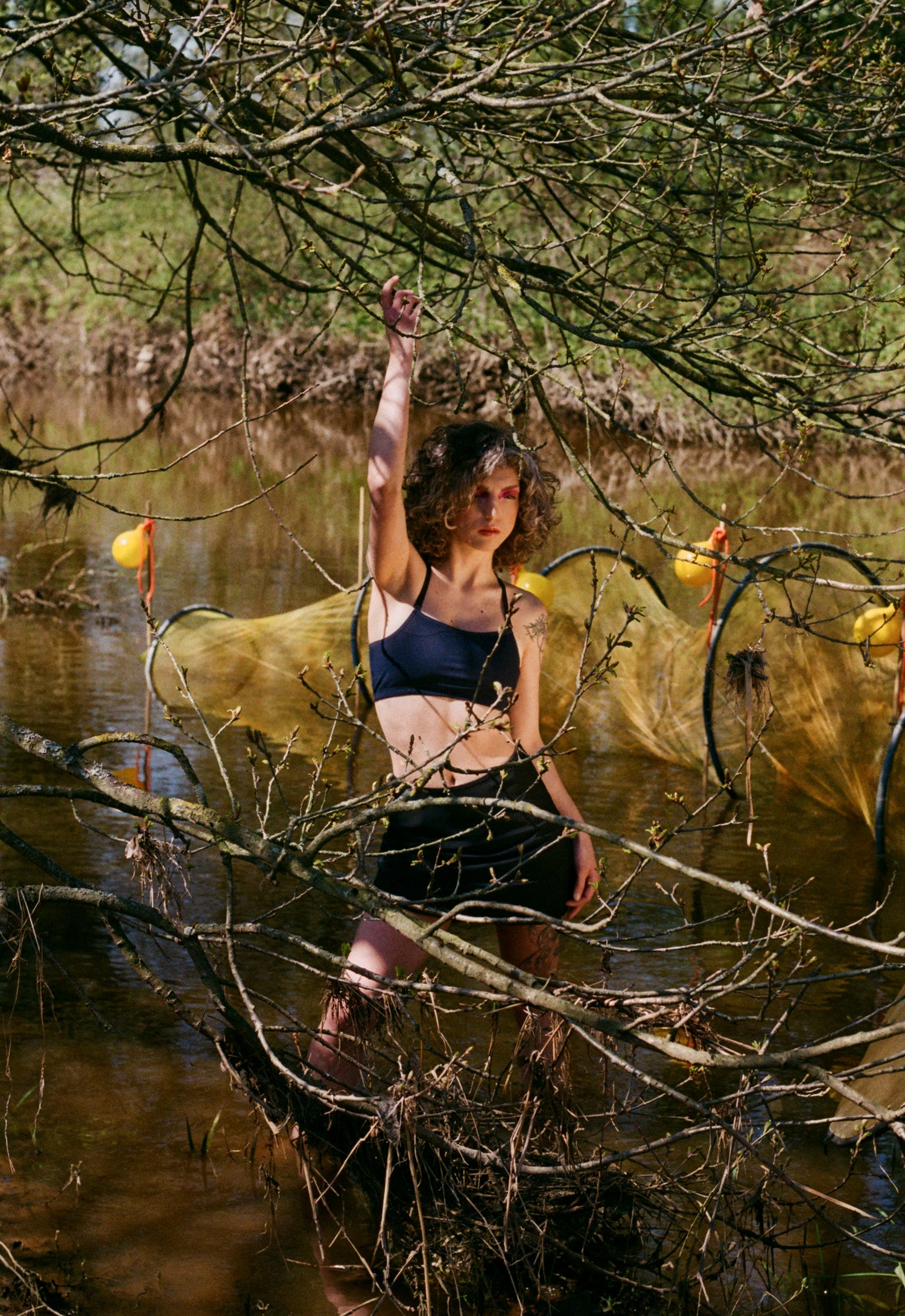 Ximena wears a prosthetic swim top and swim skirt while she poses in the water, she is holding onto branches